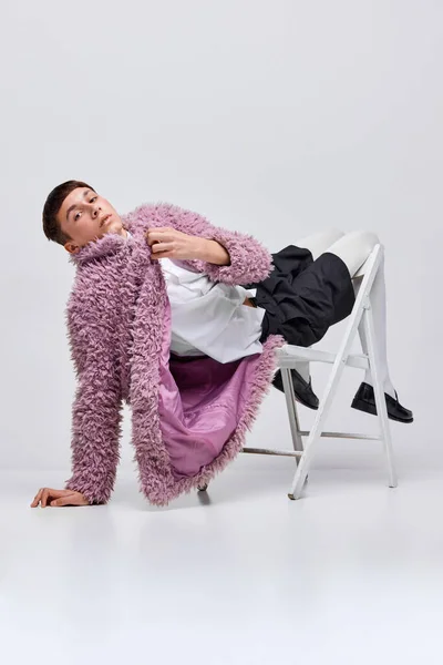 Portrait of stylish boy in pink furry coat posing on chair isolated over grey background. Extraordinary winter style. Concept of modern fashion, art photography, style, queer, uniqueness, ad