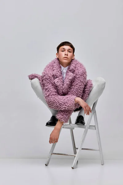 Portrait of stylish boy in pink furry coat posing on chair isolated over grey background. Youth culture. Concept of modern fashion, art photography, style, queer, uniqueness, ad
