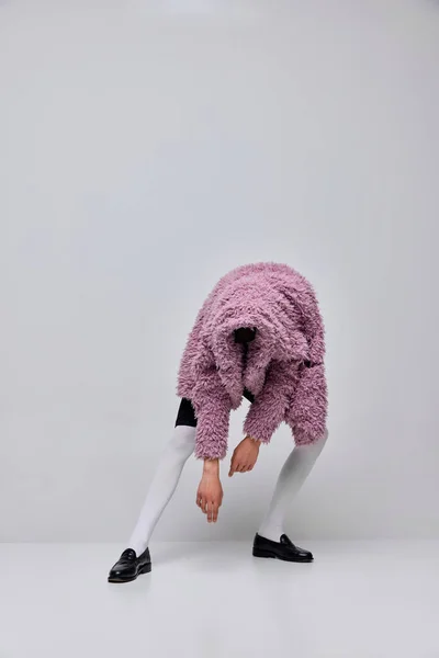 Portrait of stylish boy in pink furry coat and white tights posing isolated over grey background. Hiding face . Concept of modern fashion, art photography, style, queer, uniqueness, ad