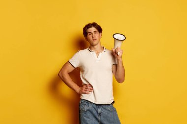 Sarı arka planda megafonla poz veren sıradan beyaz tişörtlü genç adam portresi. Haberler, satışlar. Gençlik kavramı, gündelik moda, yüz ifadesi, duygular, yaşam tarzı, reklam