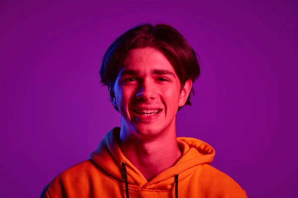 ネオンの光の中で紫の背景に隔離されたポーズカジュアルなパーカーの若い男の肖像画 カメラで微笑んだ 若者の概念 カジュアルファッション 顔の表情 ライフスタイル — ストック写真