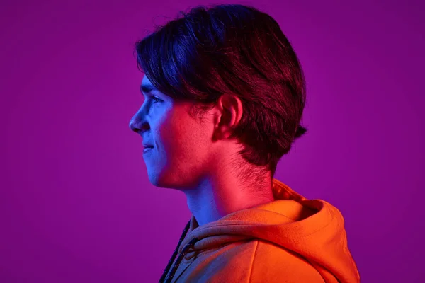 カジュアルなパーカー姿で若い男の肖像画 離れてネオンの光の中で紫の背景に孤立見て サイドビュー 若者の概念 カジュアルファッション 顔の表情 ライフスタイル — ストック写真