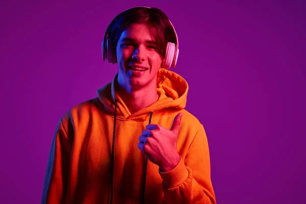 カジュアルなパーカー姿の若い男の肖像 ネオンの紫色の背景に隔離されたヘッドフォンで音楽を聴く 若者の概念 カジュアルファッション 顔の表情 ライフスタイル — ストック写真