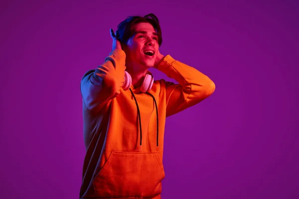 ネオンの光の中で紫色の背景に隔離されたヘッドフォンで陽気にポーズカジュアルパーカーの若い男の肖像画 若者の概念 カジュアルファッション 顔の表情 ライフスタイル — ストック写真