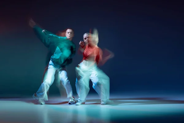 Portrait of young man and woman dancing contemp isolated over dark blue background with mixed lights. Red and green outfits. Concept of movement, youth culture, active lifestyle, action, street dance