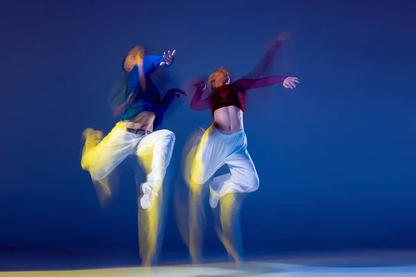 Portrait of young man and woman dancing isolated over dark blue background with mixed lights. In a jump. Concept of movement, youth culture, active lifestyle, action, street dance, ad