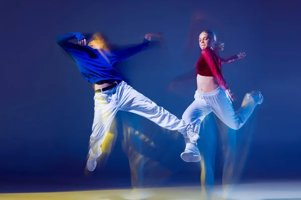Portrait of young man and woman dancing hip-hop isolated over dark blue background with mixed lights. Jumping high. Concept of movement, youth culture, active lifestyle, action, street dance, ad