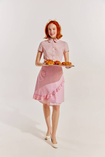 Portrait of young redhead woman in cute pink dress with freshly baked buns isolated over grey background. Concept of beauty, retro style, fashion, elegance, 60s, 70s, family. Copy space for ad