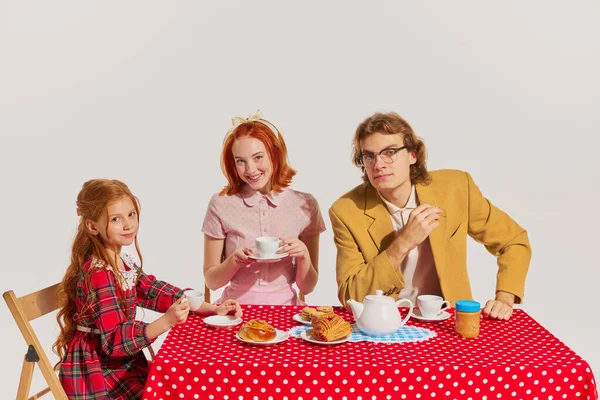 Family breakfast. Portrait of young woman, man and little girl sitting at the table isolated on grey background. Concept of beauty, retro style, fashion, elegance, 60s, 70s, family. Copy space for ad