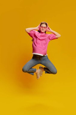 Pembe günlük tişörtlü, kulaklıkla poz veren ve sarı arka planda izole edilmiş genç bir kadın. Müzik dinliyordum. Enerji. Gençlik kültürü kavramı, duygular, yüz ifadesi, moda