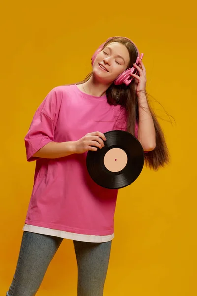 明るいピンクのTシャツの若い女性の肖像は ヘッドフォンで音楽を聞いて 黄色の背景に隔離されたポーズ ビニール音楽 若者文化 ファッションの概念 — ストック写真