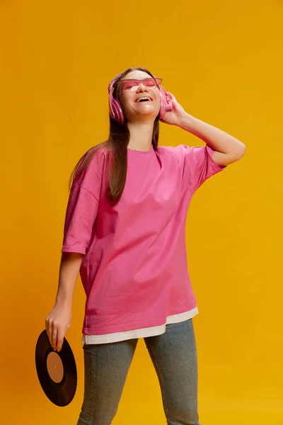Portrait of young woman in bright pink T-shirt, trendy sunglasses posing in headphones and vinyl record isolated over yellow background. Concept of youth culture, emotions, facial expression, fashion