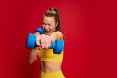 Sarı spor kıyafetli genç bir kadının portresi kırmızı arka planda izole edilmiş halterlerle egzersiz yapıyor. Motivasyon. Gençlik kültürü, duygular, yüz ifadesi, spor, yaşam tarzı, sağlık