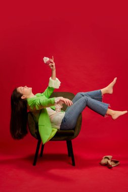 Poz veren genç bir kadının portresi, oturan ve kırmızı arka planda izole edilmiş kek yiyen. Rahatlama ve hafta sonları. Gençlik kültürü kavramı, duygular, yüz ifadesi, moda, parti