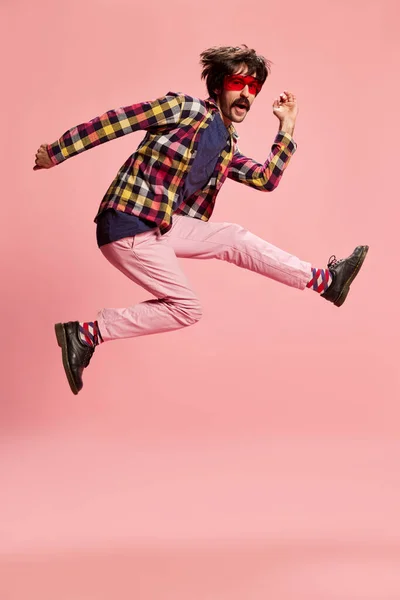 Portrait of stylish man with moustache, jumping, posing in checkered shirt and pink pants isolated over pink background. Concept of retro style, creativity, emotions, facial expression, fashion