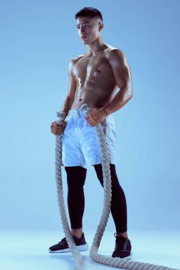 Genç kaslı adam eğitiminin portresi, mavi arka plan üzerinde izole edilmiş halatla poz veriyor. Vücut geliştirme. Spor anlayışı, fitness, sağlıklı ve aktif yaşam tarzı, motivasyon, egzersiz