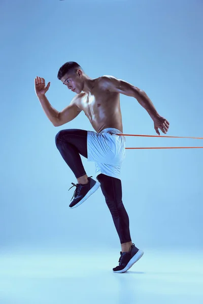 stock image Portrait of young muscular man training, running with sports expander isolated over blue background in neon light. Concept of sport, fitness, healthy and active lifestyle, motivation, strength