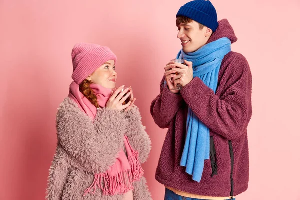 Portrait of young man and woman in knitted hat and scarf posing, warming up with mulled wine isolated over pink background. Concept of emotions, winter holidays, fashion, lifestyle, relationship