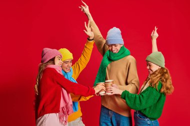 Bir grup genç, kışın giyinmiş arkadaşlar birlikte eğleniyorlar, dans ediyorlar, kırmızı arka planda izole edilmiş poz veriyorlar. Duygular kavramı, kış tatili, moda, yaşam tarzı, kutlama, arkadaşlık