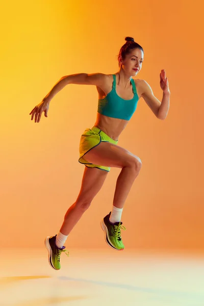 One sportive girl in sports uniform isolated over yellow background. Concept of sport, beauty, action, achievements, hobbies. Young athlete, runner looks motivated and concentrated