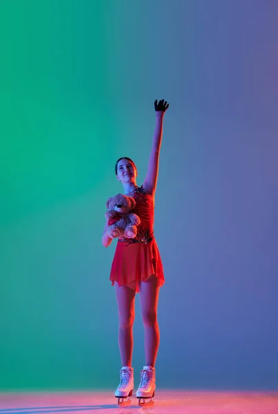 Wow, success, win. Happy young girl, female figure skater in stage costume standing with raising hand and greeting spectators of competition isolated over green-blue background in neon.