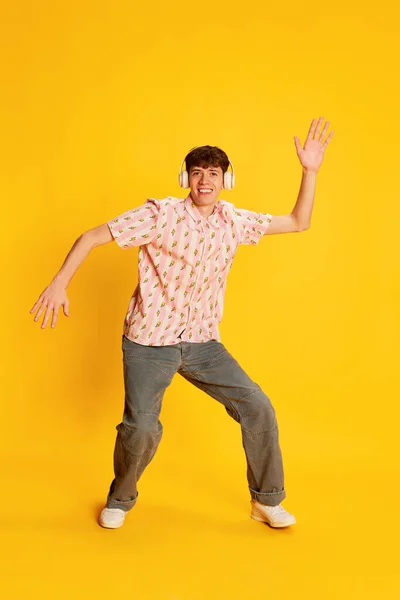 Excited young man, happy dude in vintage style clothes listening to music and dancing in headphones isolated on yellow background. Happiness, music, party, dance club and youth lifestyle concept