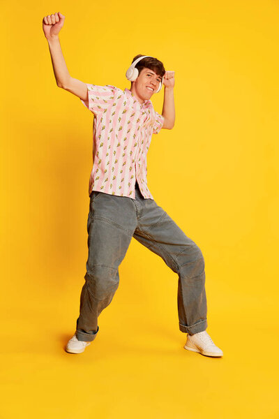 Эмоциональный улыбающийся мужчина, студент в наушниках, слушающий музыку и танцующий изолированно на ярко-желтом фоне. Счастье, музыка, вечеринка, танцевальный клуб и концепция молодежного образа жизни