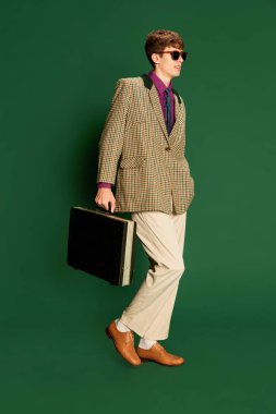 Hippi. Moda tarzı kostüm giymiş, koyu yeşil arka planda yalnız poz veren genç bir adam. Retro biçim, yaratıcılık, duygular, yüz ifadesi, moda ve iş dünyası kavramı