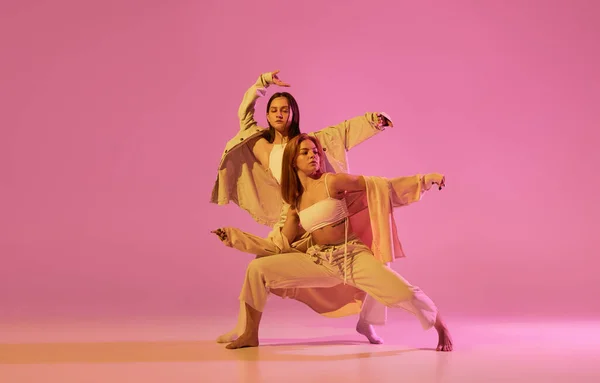 Modern dance art. Young girls, couple of dancers in sports style clothes dancing experimental dances isolated over light pink background in neon. Concept of music, emotions, dance. Copy space for ad