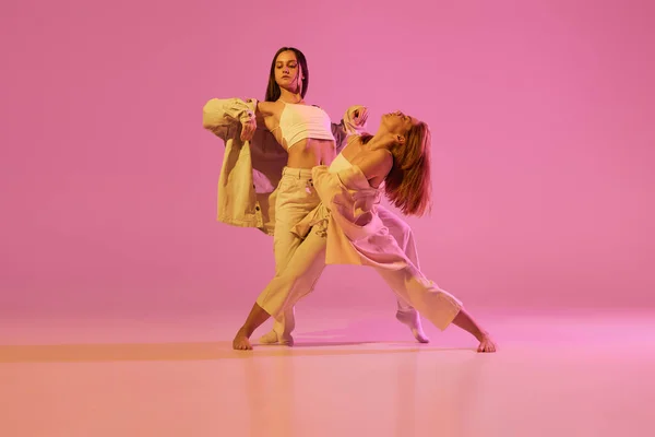 Modern dance art. Young girls, couple of dancers in sports style clothes dancing experimental dances isolated over light pink background in neon. Concept of music, emotions, dance. Copy space for ad
