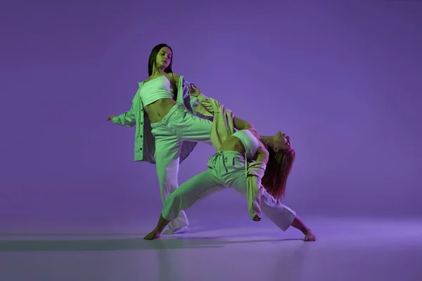 Experimental dance. Two young girl in motion, action isolated over purple background. Concept of new dance style, youth culture, music and fashion, ad, trends. Flexible and graceful dancers