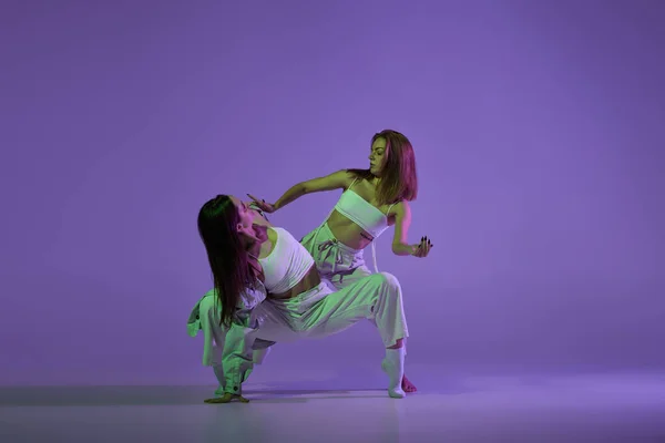 Experimental dance. Two young girl in motion, action isolated over purple background. Concept of new dance style, youth culture, music and fashion, ad, trends. Flexible and graceful dancers