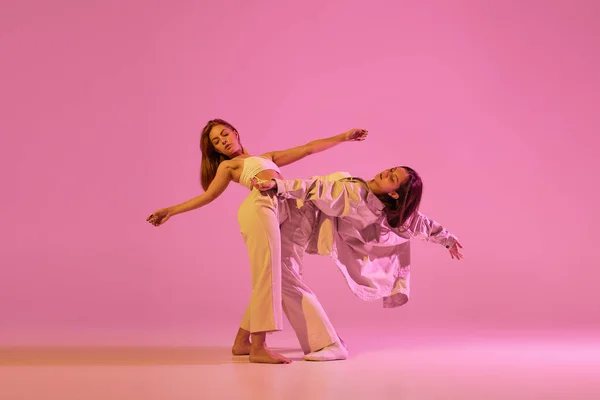 Love. Young girls, couple of dancers in sports style clothes dancing experimental dances isolated over light pink background in neon. Concept of music, emotions, dance. Copy space for ad