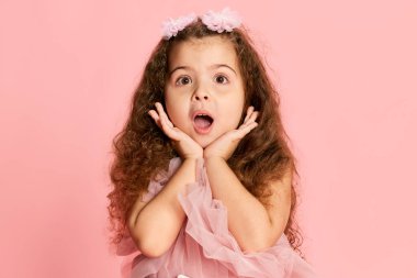 Şaşkın şirin küçük kız, uzun kıvırcık saçlı çocuk pembe arka planda soyutlanmış prenses elbisesi giyiyor. Mutlu tasasız çocukluk, çocuk duyguları, çocuk modası ve reklam. Mutlu Noeller.