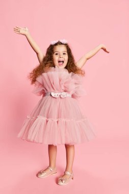 Zevk. Çekici, küçük bir kızın portresi, pembe arka planda izole edilmiş uzun kıvırcık saçlı bir çocuk. Mutlu çocukluk, çocuk duyguları, moda ve reklam. Mutlu görünüyor, mutlu.