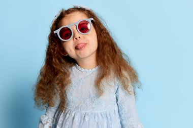 Aptal ruh hali. Mavi elbiseli okul öncesi kızın, mavi arka planda izole edilmiş olumlu duygularını ifade ettiği bir fotoğraf. Yüz ifadeleri, reklam, mutluluk, çocuk modası ve reklam konsepti. Tek Renkli