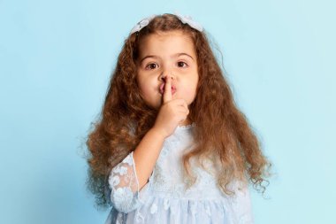 Sırlar. Mavi elbiseli anaokulu çocuğunun stüdyosu mavi arka planda izole edilmiş parmağını dudaklarına bastırıyor. Yüz ifadeleri, reklam, mutluluk, çocuk modası ve reklam konsepti. Tek renkli portre