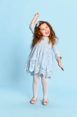 Eğlence, neşe. Mavi elbiseli neşeli anaokulu kızı mavi arka planda izole edilmiş pozitif duygularını ifade ediyor. Yüz ifadeleri, reklam, mutluluk, çocuk modası ve reklam konsepti. Tek renkli portre