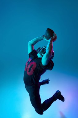 Atla! Amerikan futbolcusunun spor üniformalı ve koruyucu kasklı görüntüsü mavi arka planda, neon ışıkta izole edilmiş. Profesyonel spor, takım, yarışma, şampiyonluk kavramı
