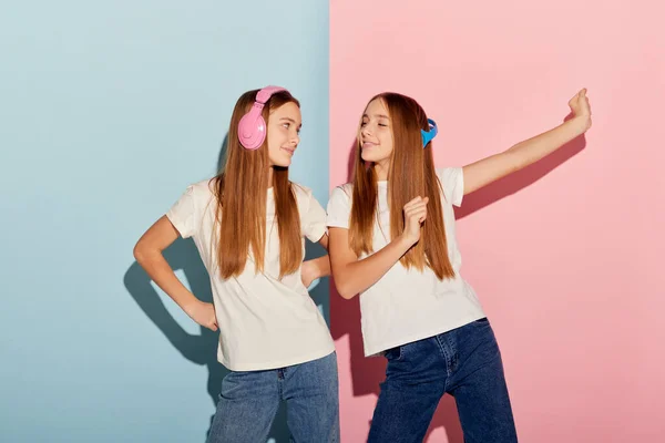 Dance. Charming young girls, two sisters friends in casual style clothes isolated over pink-blue studio background. Concept of youth fashion, relationship, emotions, beauty and music.