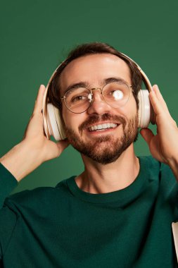 Sakallı, sıradan süveter giyen ve yeşil arka planda gözlük takan yakışıklı bir adamın yakın plan portresi. Mutluluk, olumlu duygular. Tek Renkli