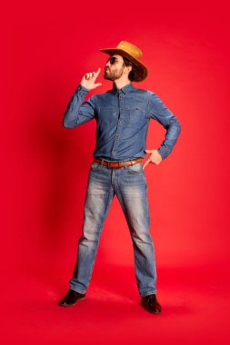 Kovboy kıyafetli heyecanlı genç adam eğleniyor, kırmızı arka planda yalnız poz veriyor. Kovboy şapkası ve kot pantolon giymiş bir model. Moda, duygu konsepti. Mutlu görünüyor, mutlu.