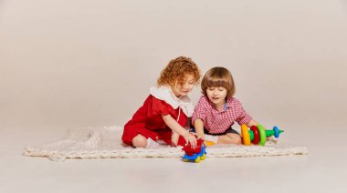 İki sevimli çocuk, kız kardeş ve erkek kardeş yerde oturup gri arka planda izole bir şekilde oyun oynuyorlar. Mutlu çocukluk kavramı, boş zaman aktiviteleri, eğlence, yaşam tarzı, aile, eski tarz