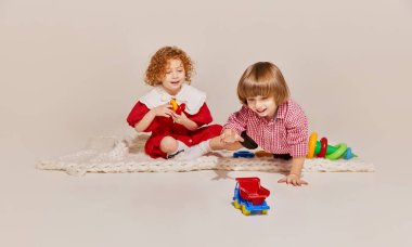 İki sevimli çocuk, kız kardeş ve erkek kardeş yerde oturup gri arka planda izole bir şekilde oyun oynuyorlar. Mutlu çocukluk kavramı, boş zaman aktiviteleri, eğlence, yaşam tarzı, aile, eski tarz