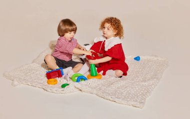 İki sevimli çocuk, kız kardeş ve erkek kardeş yerde oturup gri arka planda izole bir şekilde oyun oynuyorlar. Mutlu çocukluk kavramı, boş zaman aktiviteleri, eğlence, yaşam tarzı, aile, eski tarz