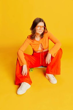 Turuncu bluzlu, kaykaylı, sarı arka planda pantolonu olan pozitif bir kız. Gençlik, moda, aktif yaşam tarzı, mutlu duygular ve aktif yaşam tarzı konsepti. İlan için alanı kopyala