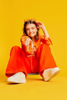 Duygusal açıdan güzel bir kız, turuncu bluzlu ve sarı arka planda kaykaylı pantolonu var. Gençlik, moda, aktif yaşam tarzı, mutlu duygular ve aktif yaşam tarzı konsepti. İlan için alanı kopyala