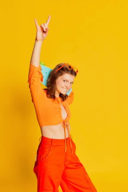 Renkli sarı arka planda kaykaylı turuncu bluzlu ve pantolonlu heyecanlı kız. Gençlik, moda, aktif yaşam tarzı, mutlu duygular ve aktif yaşam tarzı konsepti. İlan için alanı kopyala