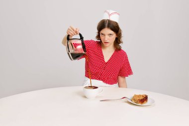 Stylish young female waitress in retro american fashion style of 70s, 80s sitting at table and drinking coffee over grey background. Funny meme emotions, ad, sales. Weird people concept