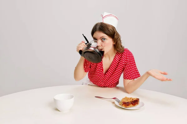 Stylish young female waitress in retro american fashion style of 70s, 80s sitting at table and drinking coffee over grey background. Funny meme emotions, ad, sales. Weird people concept
